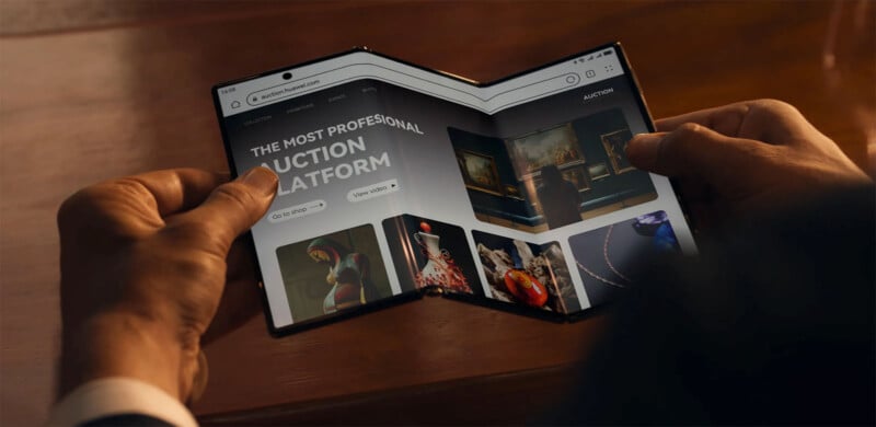 A person holding a flexible foldable tablet opened to an online auction platform. The screen shows various categories including paintings, sculptures, and jewelry. The tablet has a noticeable bend in the middle, demonstrating its flexibility.