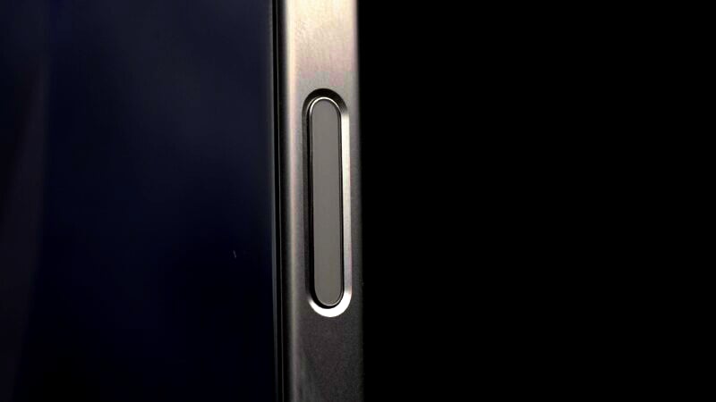 Close-up of the side of a smartphone showing a metallic volume button. The button is elongated with rounded edges and sits within a glossy, dark-colored frame. The right side of the frame is visible against a black background, while the left side edge appears slightly reflective.