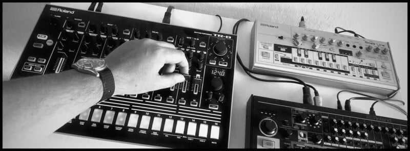 A black and white image of a hand adjusting knobs on a Roland TR-8 drum machine. Other electronic music equipment, including a Roland TB-03 and Roland TR-09, are connected and positioned around the main device. The person is wearing a wristwatch.