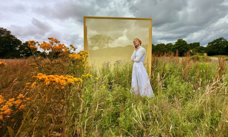 A woman in a white dress stands in a field of tall grass and yellow flowers, with a large rectangular golden frame behind her. The sky above is cloudy, creating a dramatic backdrop. The scene has a serene and contemplative atmosphere.