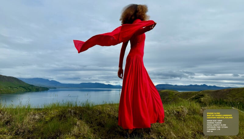 A person in a long, flowing red dress stands on a grassy hill overlooking a serene lake and mountains in the background. The dress and scarf billow in the wind. The sky is overcast with a mix of clouds.