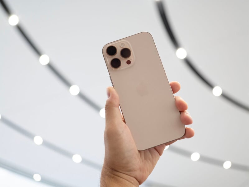 A hand holding a gold-colored iPhone with three camera lenses on the back. The background features a modern, curved, brightly-lit ceiling with circular lights.