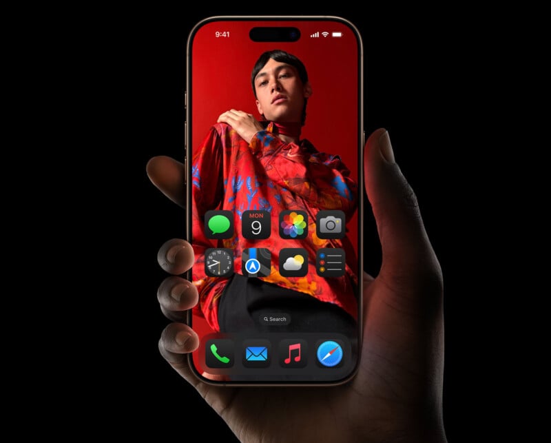 A hand holding a smartphone displaying a home screen with various app icons. The background of the screen features a person wearing a vividly colored, red and blue patterned shirt, posing against a red backdrop. The image is set against a black background.