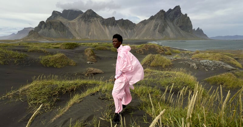 A person wearing a light pink outfit stands on a dark, sandy beach with patches of tall grass. In the background, there are dramatic, rugged mountains and a cloudy sky. The person looks to the side, with the landscape stretching out behind them.