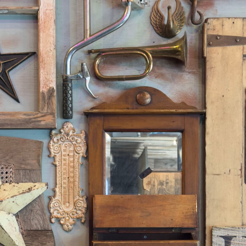 A wooden wall display featuring rustic decorations, including a vintage bugle, a plumbing pipe handle, ornate wall plaques, and a mirror with a wooden frame. Various textures and items create a nostalgic, antique atmosphere.