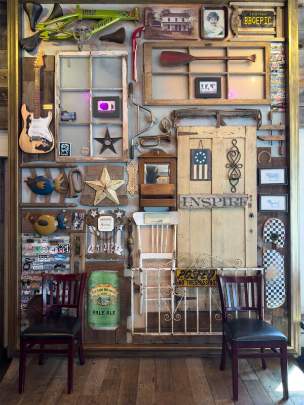 A wall decorated with a mix of vintage items including a guitar, trumpet, old windows, a door, various pictures, signs, skateboards, and other knick-knacks. Two wooden chairs with black seats are placed against the wall on a wooden floor.