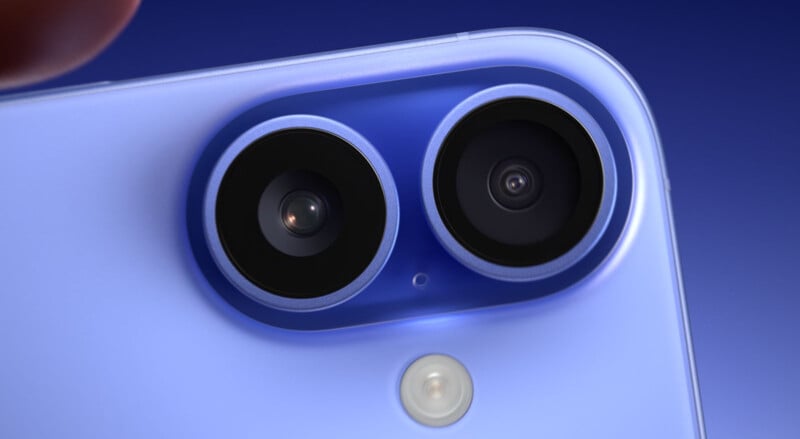 Close-up view of the back of a smartphone with a dual camera setup. The lenses are encircled in a slightly raised oval module, accompanied by a flash positioned below. The phone has a smooth, light purple finish against a dark blue gradient background.