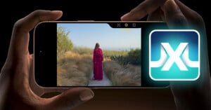 A person holds a smartphone horizontally with an image of a person in a red dress standing on a wooden path in a grassy outdoor area displayed on the screen. There is also a glowing icon featuring an "X" on the right side of the image.