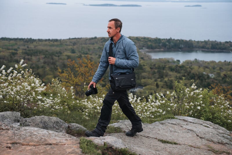 A person in outdoor attire walks on a rocky path, holding a camera and carrying a shoulder bag. Behind them, there is a scenic backdrop of a forested area with a body of water and islands in the distance. White flowers and greenery are in the foreground.