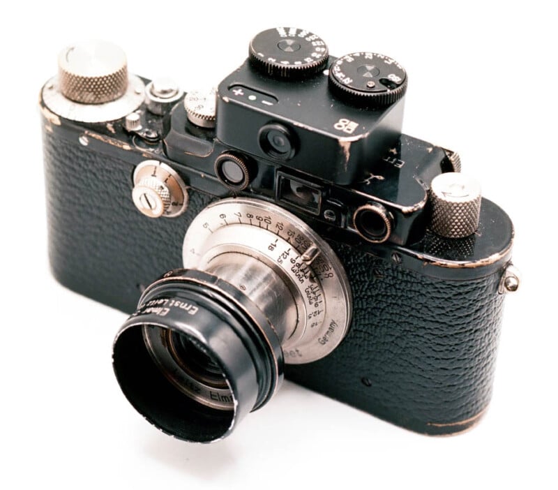 A vintage black Leica camera with a metal lens and several intricate dials. The body shows signs of wear, giving it a classic, aged appearance. The lens has "Germany" and "Elmar" markings. The camera is positioned on a white background.