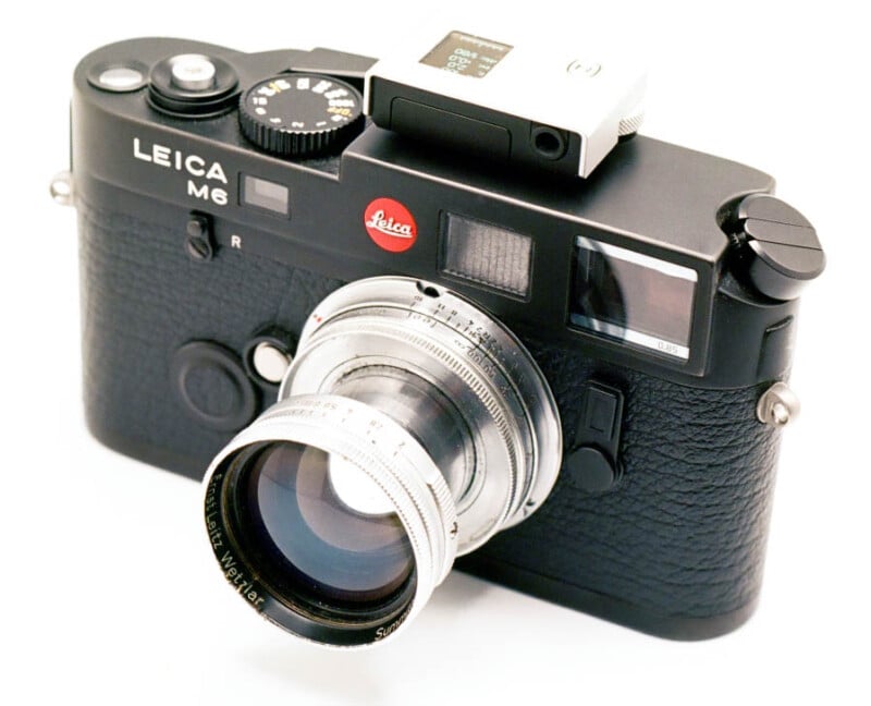 A black Leica M6 35mm rangefinder film camera is shown, equipped with a silver lens. The camera has the Leica red logo on the front and a mounted external accessory on top. The body features classic controls and a sleek, compact design against a white background.