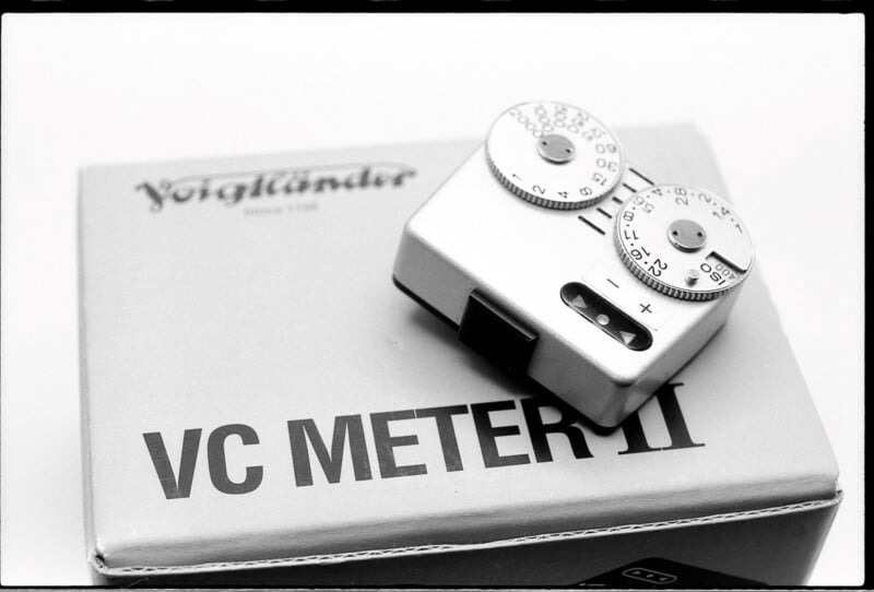 A Voigtländer VC Meter II sits atop its gray packaging box. The light meter features two dials on its top side, used for adjusting and reading exposure settings. The Voigtländer logo is partially visible on the box. The image is in black and white.