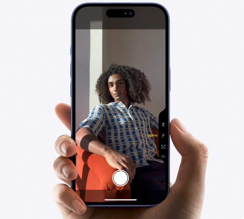 A hand is holding a smartphone, displaying a camera viewfinder. The screen shows a well-lit person with curly hair, wearing a patterned shirt, seated on a red chair. The person's face is turned slightly to the side, and the background appears light and minimalistic.