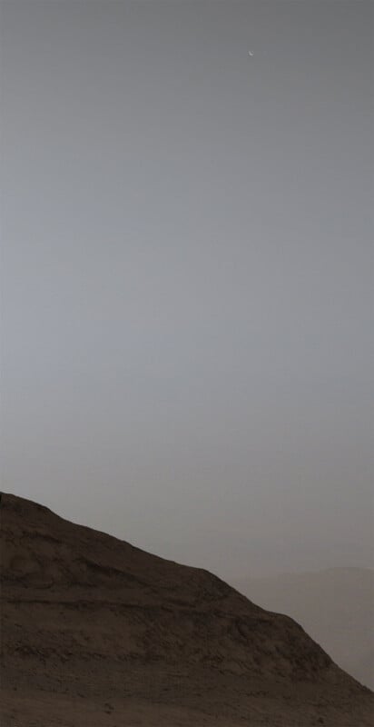 A dusk scene with a rocky, barren landscape in the foreground and a clear, dimly lit sky above. A slender crescent moon and a tiny distant planet are visible high in the sky, adding a celestial element to the stark, quiet scenery below.