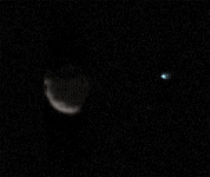 A dark, grainy image showing a faintly illuminated crescent-shaped object on the left side and a small, bright, blueish speck of light on the right side against a black background.