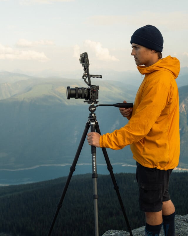 A person in a bright yellow jacket and black beanie hat stands on a mountain edge, using a camera on a tripod. The person is focused on the camera controls. The background shows expansive, scenic mountain ranges under a partly cloudy sky.