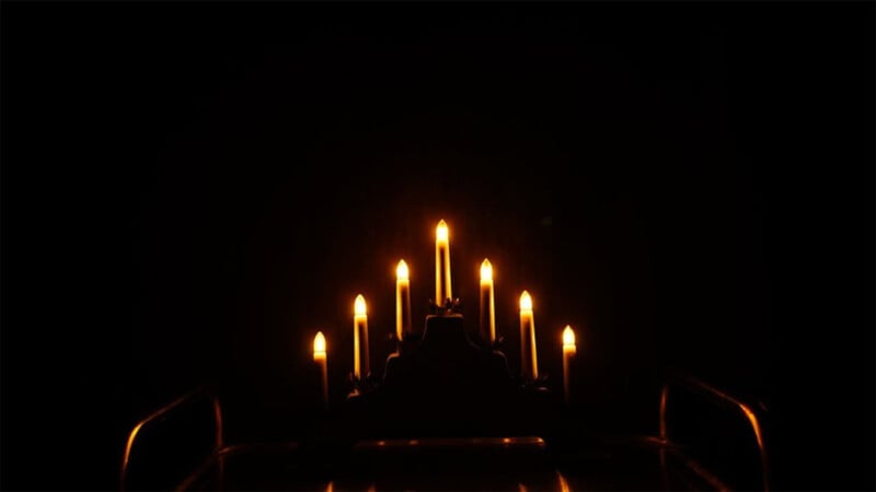 A dark background highlights seven lit candles arranged in a candelabra, each flame glowing warmly and casting a soft light. The candelabra's base is partially visible, rising from the bottom of the image. The scene evokes a sense of calm and serenity.