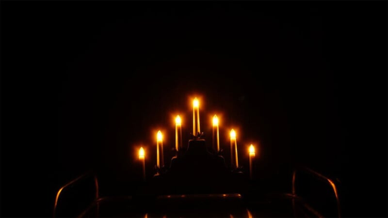 A group of seven lit candles arranged in a candelabrum in a dark setting. The candles emit a warm, flickering light that illuminates their immediate surroundings while the background remains pitch black.