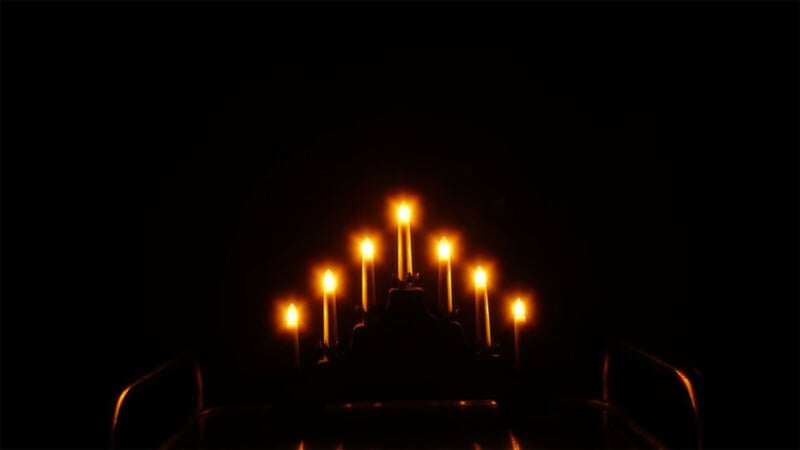 Dimly lit chandelier with seven glowing candles against a dark background, creating a warm and moody atmosphere. The candles are arranged in a symmetrical pattern, casting a soft, yellowish light.