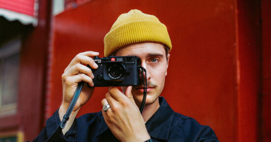 A person wearing a yellow beanie and a dark jacket holds a camera up to their face, looking directly into the lens. The background features a red wall creating a vibrant, contrasting backdrop.