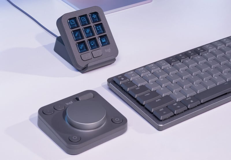 A modern workspace setup features a multi-button input device with nine programmable buttons on a stand, a sleek grey keyboard with keycaps, and a separate compact control dial with additional buttons, all placed on a white desk.