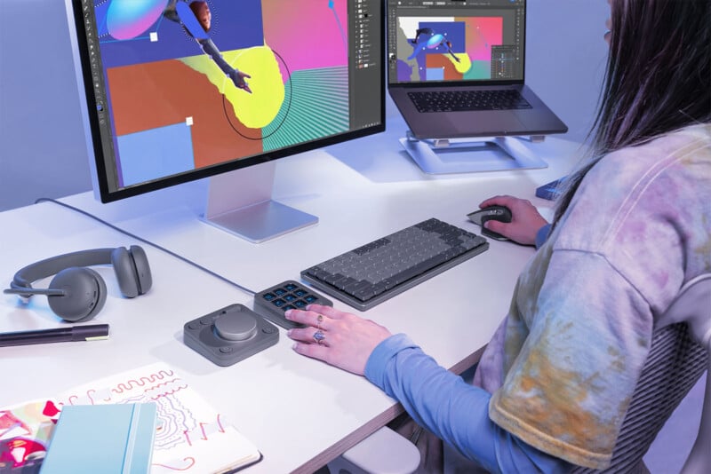 A person in a tie-dye shirt sits at a desk using a computer with a graphics tablet and stylus. The monitor displays colorful digital art. The desk also has a mechanical keyboard, headphones, a notebook, a pen, and a custom editing controller.