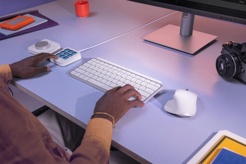 A person with dark skin, wearing a brown long-sleeve shirt with a bracelet, uses a white keyboard and mouse on a cluttered desk. On the desk are a large monitor, an orange external hard drive, a camera, a cup, and a keypad with multiple blue buttons.