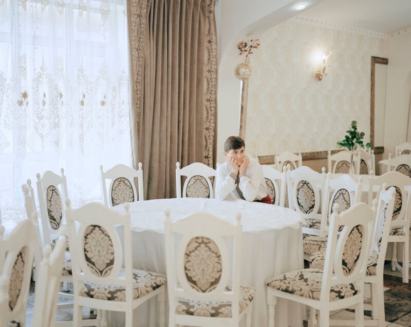 A person in formal attire sits alone at a round table in an elegantly decorated room, surrounded by empty chairs. The space features ornate curtains, floral-patterned upholstery on chairs, and soft, ambient lighting.