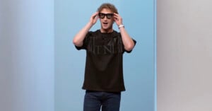 A person with curly hair wearing a black T-shirt, jeans, and large black glasses stands against a blue and white background, smiling with hands touching the glasses.