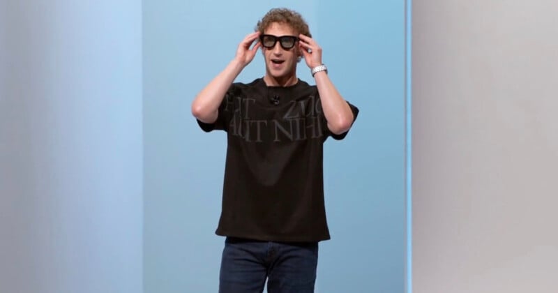 A person with curly hair wearing a black T-shirt, jeans, and large black glasses stands against a blue and white background, smiling with hands touching the glasses.