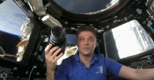 An astronaut inside a spacecraft holds a camera, with Earth visible through the windows in the background. He wears a blue polo shirt and is surrounded by various equipment and controls. The scene is well-lit, showcasing the interior details of the spacecraft.