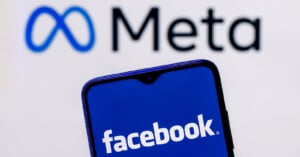 A smartphone displaying the Facebook logo is held in front of a blurred Meta logo background.