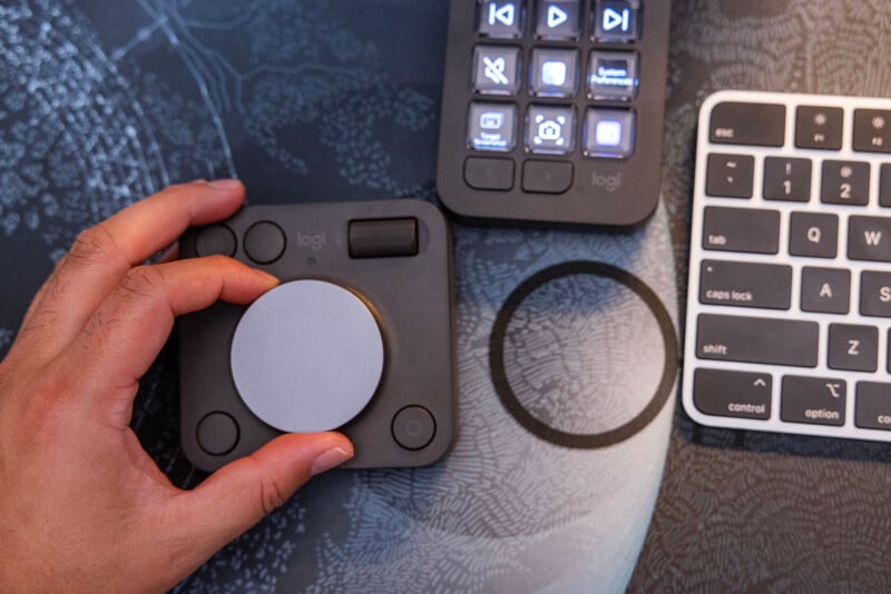 A person adjusts the dial on a Logitech control device placed on a workspace with a textured mat. Other gadgets, including a Logitech button panel and a partial view of a white keyboard, are also present. The workspace appears organized and tech-focused.