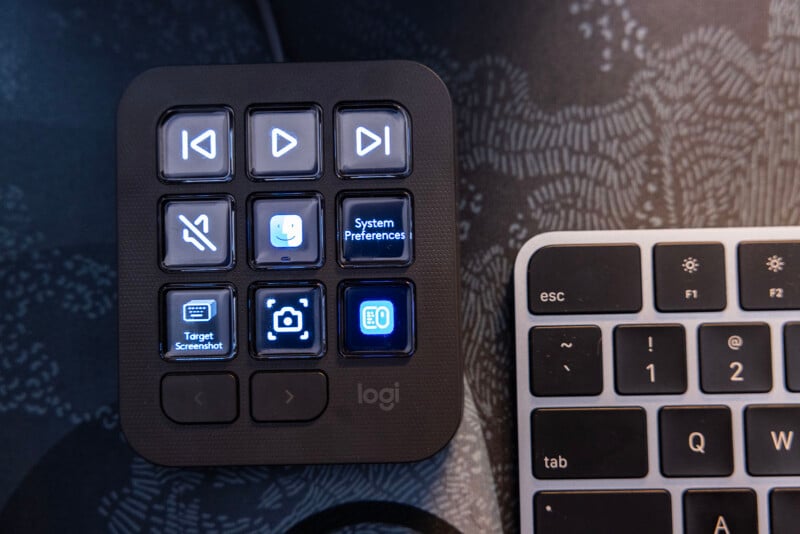 A close-up of a small, black control pad with various illuminated buttons, including playback controls, system preferences, mute, camera, and screenshot icons. It is positioned next to a portion of a keyboard, showing the "esc" and function keys.