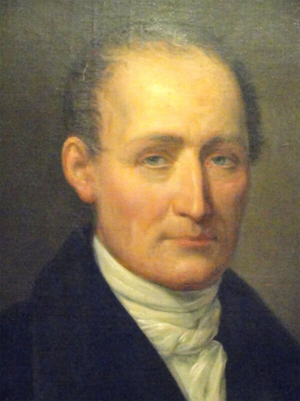 A portrait of an older man with a receding hairline and somber expression, dressed in early 19th-century fashion. He wears a dark coat and a white cravat. The background is plain and dark, drawing focus to his thoughtful facial features.