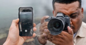 Close-up image of a person taking a photo with a Nikon DSLR camera on the right side. A hand is holding a smartphone with its camera app open on the left side, showing the same scene. The background is an outdoor, slightly misty setting.