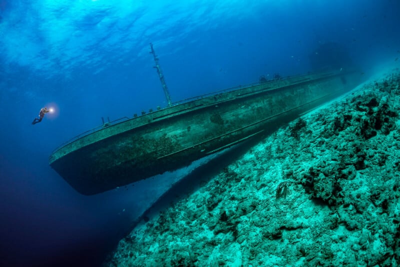 A scuba diver explores a large, sunken ship resting on its side on the ocean floor. The ship appears rusty and aged, with marine life surrounding it. The diver holds a flashlight, illuminating a small area of the ship's structure. The water is clear and blue.