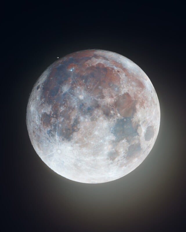 A high-resolution image of the full moon against a dark sky. The lunar surface displays vibrant details including various shades of grey, white, and hints of red and blue. Craters, maria, and highlands are clearly visible, highlighting the moon's diverse topography.