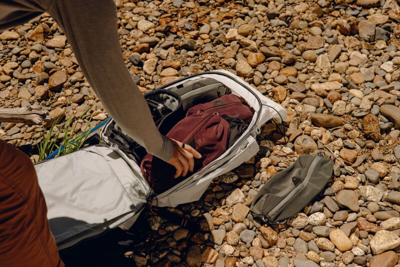 A person in a long-sleeve shirt packs or unpacks a white backpack placed on a rocky ground, with a brown jacket inside. A small gray pouch lies beside the backpack. The surroundings suggest an outdoor or hiking setting.