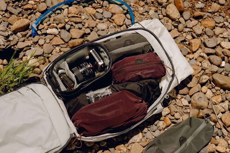 A white travel backpack lies open on a rocky surface, revealing organized compartments filled with camera gear, including a camera with a lens, additional lenses, and various cases and pouches. A blue hydration tube is partially visible in the background.