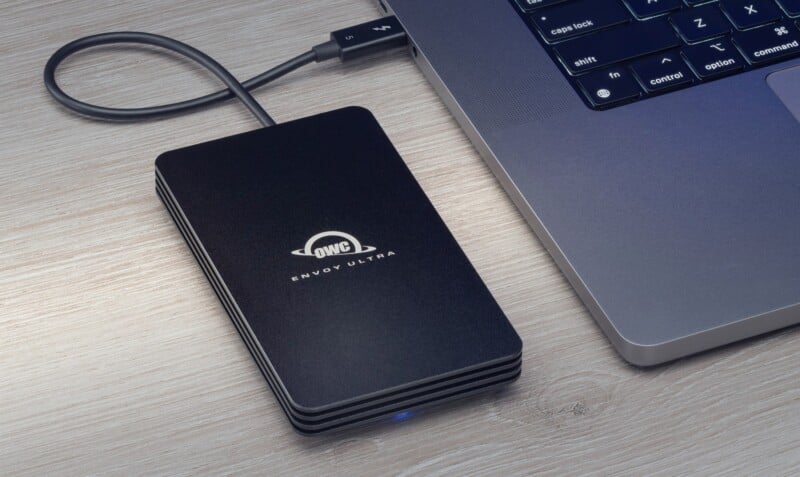 A black OWC Envoy Ultra external SSD is connected to a laptop via a USB cable. The devices are placed on a wooden surface, and the laptop keyboard is partially visible in the corner of the image.