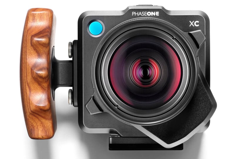 Close-up of a PhaseOne XC professional camera. The camera features a sleek metallic body with a large, protruding lens and a wooden handle on the left side. A blue button appears next to the lens, and a lens hood is attached for light protection.