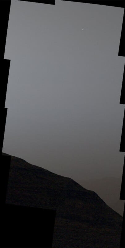 A hazy sky with a faint crescent shape near the top, likely the Moon or a planet, above a dark, rugged landscape in the foreground. The image has a patchy appearance from being a mosaic of multiple photographs.