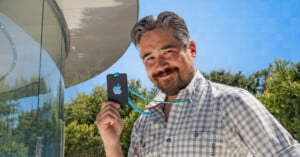 A smiling man with short gray hair and a beard holds up an Apple logo ID badge on a lanyard. He is wearing a checkered shirt and stands in front of a modern glass building with trees in the background under a clear blue sky.