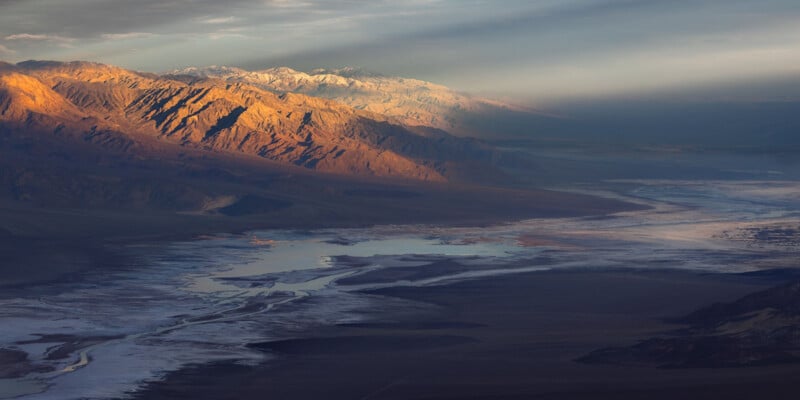 A vast desert landscape at sunset, with rugged mountains partially covered in snow. The sun casts a warm, golden light on the mountain ridges and valleys below, while patches of shadowed areas and a reflective body of water contrast with the illuminated peaks.