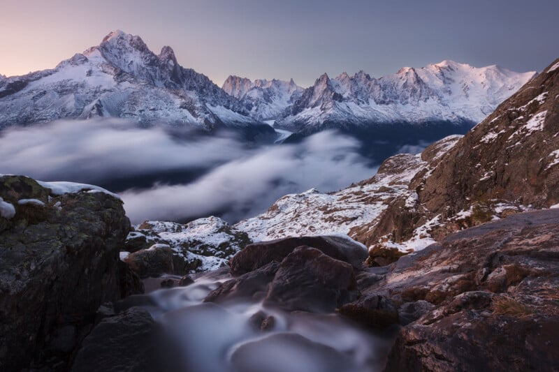 A breathtaking mountain landscape featuring snow-capped peaks under a clear sky at dusk. A river with a silky water effect flows over rocks in the foreground, while low clouds linger in a valley beneath the majestic mountains in the background.