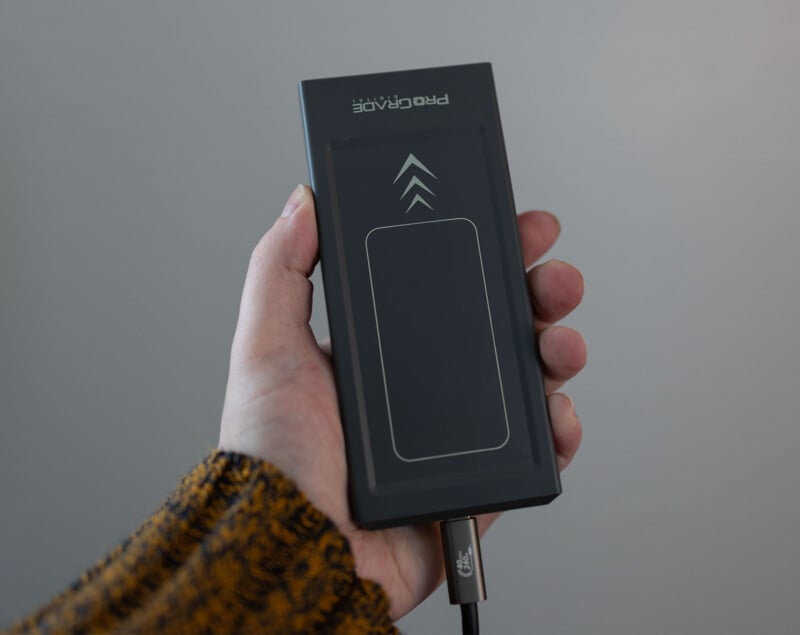 A hand holding a black rectangular wireless charger labeled "PRO CHARGE." The charger has a simple design with a rectangular outline and two upward-pointing arrows on its surface. A charging cable is connected at the bottom. The person is wearing a mustard-colored sweater.