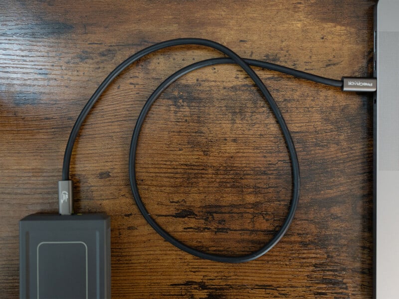 A black external hard drive is connected to a laptop with a USB-C cable. The laptop is partially visible on the right side of the image, and the hard drive is on the left. The connection cable arcs in a loose loop on a wooden surface.