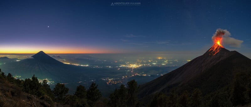 A panoramic nighttime view of an erupting volcano with lava and smoke on the right. Another, dormant volcano stands on the left. City lights speckle the landscape below, and the sky transitions from twilight on the horizon to starry night above.