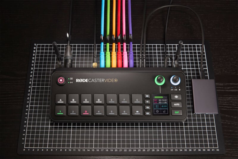 A RØDECaster Video device placed on a gridded surface. The device has multiple colored cables connected to the top and various buttons, knobs, and a small display screen on the front panel. The black casing has labels and an OLED screen showing status information.